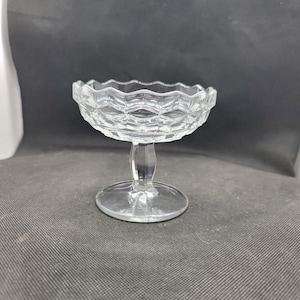 Puede incluir: Un cuenco de postre de cristal transparente, con borde festoneado y diseño de diamantes. Se apoya sobre un tallo corto y una base redonda. El cristal es transparente, permitiendo el paso de la luz.