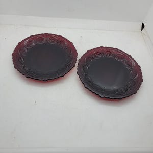May include: Two ruby red glass plates with a scalloped edge and a detailed pattern. The plates have a deep, rich color and a classic design, suitable for serving appetizers or desserts. The plates are likely vintage or antique.