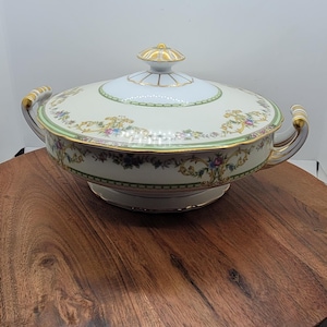 Może przedstawiać: Przykryta misa do serwowania z uchwytami, z kremową podstawą i pokrywką. Misa jest ozdobiona kwiatowymi i złotymi akcentami i spoczywa na drewnianej powierzchni. Misa ma średnicę około 30 cm.