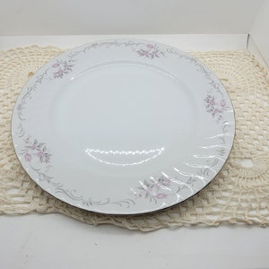 May include: White dinner plate with a gold rim and a delicate floral and scroll design in pink and gray. The plate is part of a set and is displayed on a cream-colored lace doily.