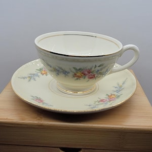 May include: Cream-colored teacup and saucer set with floral patterns in pink, yellow, and blue. The cup and saucer have gold trim. The set is placed on a wooden surface, suggesting a vintage or antique aesthetic.