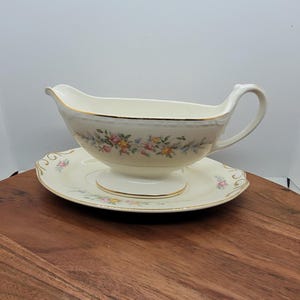 May include: Cream-coloured gravy boat and saucer set with floral and gold trim. The gravy boat has a handle and a spout. The saucer is round. The set is on a wooden surface. A classic addition to any dining table.