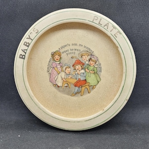 Puede incluir: Un plato de bebé de cerámica blanca con un borde verde. El plato presenta una ilustración colorida de niños tocando música. El texto en el plato dice "BABY'S PLATE" y "Tom.hewas a piper's son. He learned when he was young to play".