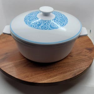 Puede incluir: Cazuela de cerámica blanca con tapa con diseño floral azul. El plato tiene dos asas laterales y un pomo blanco en la tapa. Se encuentra sobre una bandeja de madera.