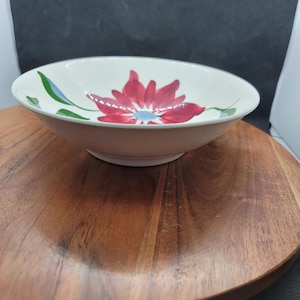 May include: A cream-colored ceramic bowl with a vibrant red floral design at the center. The flower has a blue center and is surrounded by green leaves. The bowl is set on a wooden surface.