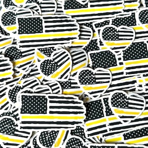 May include: A pile of thin black and yellow American flag stickers with a thin yellow stripe across the middle. Some of the stickers are in the shape of a heart.