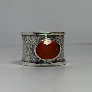 May include: A silver ring with a large, round, orange gemstone set in the center. The band of the ring is decorated with a repeating pattern of small flowers.