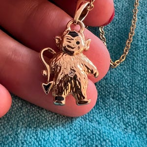 Könnte beinhalten: Goldfarbene Halskette mit einem kleinen, detaillierten Teufelspannchen. Das Anhänger hat ein schwarzes Gesicht mit einem weißen Auge und einen kleinen Diamanten in der Mitte seiner Brust.