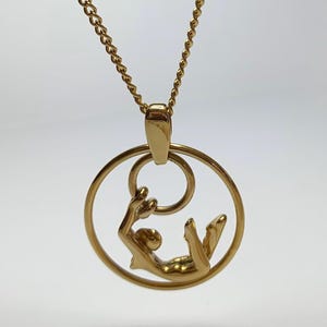 May include: Gold-toned pendant necklace featuring a stylized figure within a circular frame. The figure is depicted in a reclining pose, with a smaller ring above. The chain is a delicate, linked design.