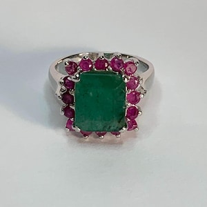 Op de afbeelding: Een zilveren ring met een rechthoekige smaragd als middensteen, omringd door een halo van kleine, ronde robijnen. De ring heeft een gepolijst, reflecterend oppervlak, tegen een witte achtergrond.