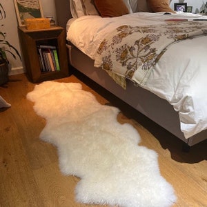May include: A white faux fur rug with a shaggy texture, placed on a wooden floor next to a bed with a white duvet and a floral patterned throw.
