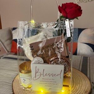 May include: A clear tote bag filled with gifts, including a candle, a notebook, a compact mirror, and a decorative block with the word "Blessed." A red rose and string lights adorn the bag. The tote sits on a wooden slice.