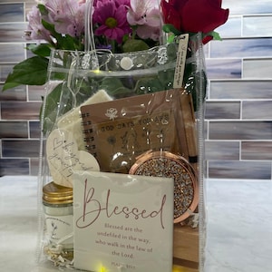 May include: A transparent tote bag containing a variety of items, such as a notebook, a compact mirror, a candle, and a card with the word "Blessed". The bag also features a red rose and a bouquet of pink and purple flowers. The text "GOD SAYS YOU ARE" is visible.