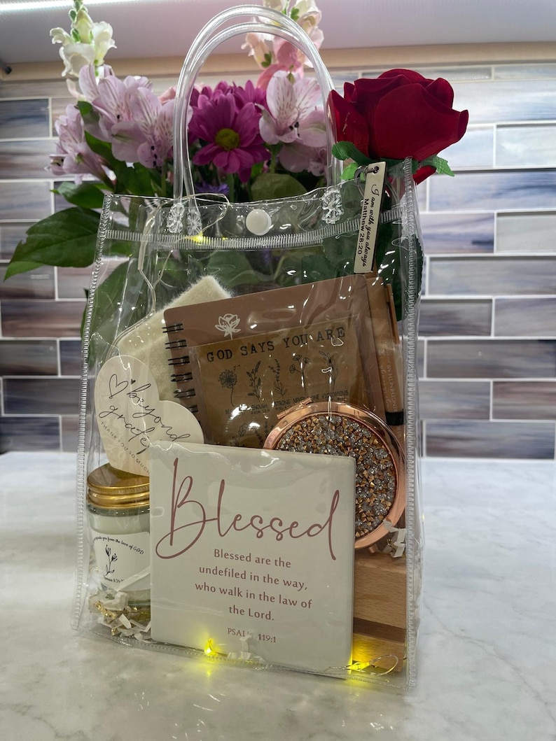 May include: A transparent tote bag containing a variety of items, such as a notebook, a compact mirror, a candle, and a card with the word "Blessed". The bag also features a red rose and a bouquet of pink and purple flowers. The text "GOD SAYS YOU ARE" is visible.