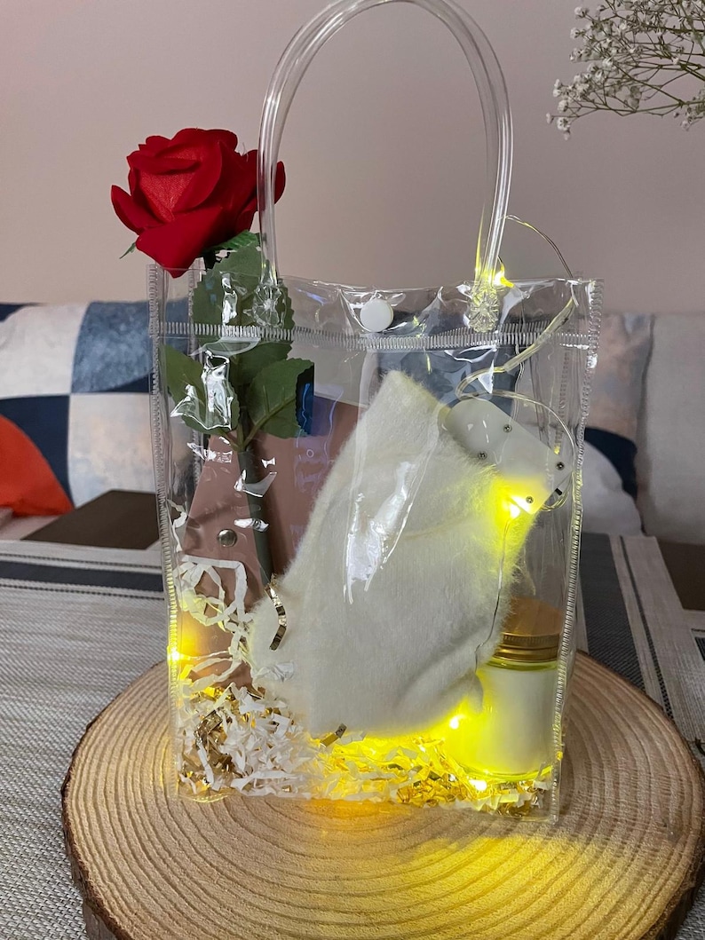 May include: A transparent tote bag containing a red rose, a white plush item, a small jar, and decorative filler. The bag is lit by yellow fairy lights and rests on a wooden slice.