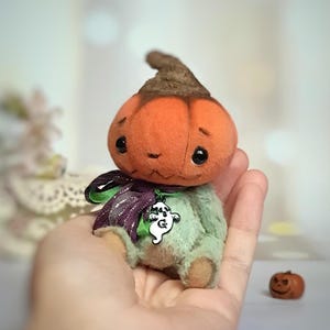 May include: A plush pumpkin doll with an orange head and a green body. The doll has a brown stem, black eyes, and a purple and green ribbon around its neck, with a ghost charm. A small pumpkin is in the background.