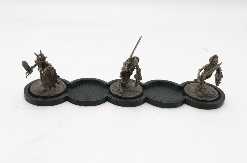May include: Three miniature skeletal figures with swords and shields are displayed on a black plastic base with three circular indentations.