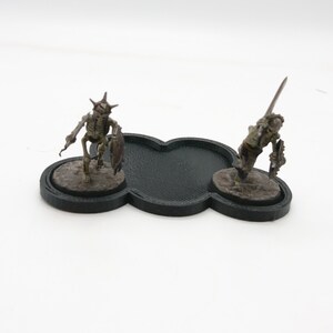 May include: Two miniature skeletal figures with swords and shields stand on black circular bases. The bases are connected by a black, cloud-shaped piece.
