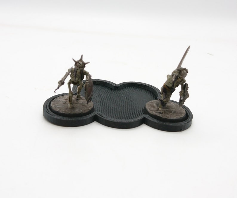 May include: Two miniature skeletal figures with swords and shields stand on black circular bases. The bases are connected by a black, cloud-shaped piece.