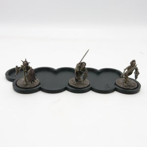 May include: Three miniature skeletal figures with swords and shields stand on a black cloud-shaped base. The figures are painted in shades of brown and green.