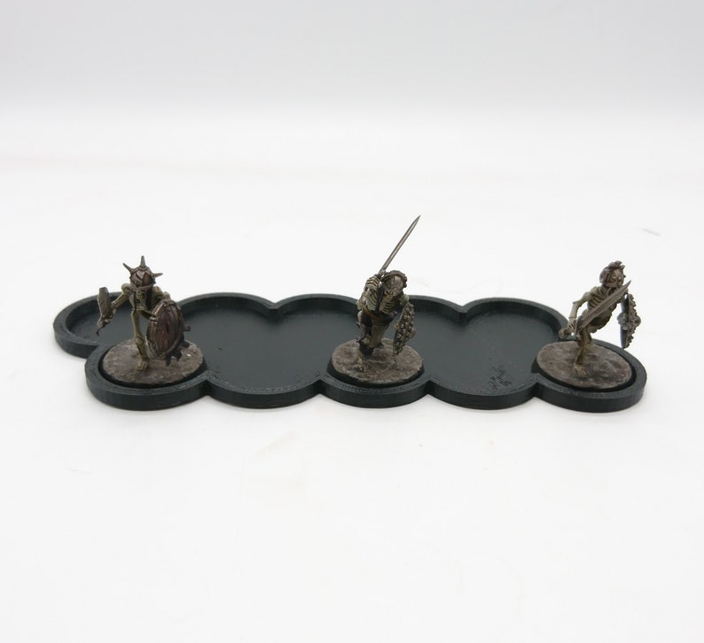 May include: Three miniature skeletal figures with swords and shields stand on a black cloud-shaped base. The figures are painted in shades of brown and green.