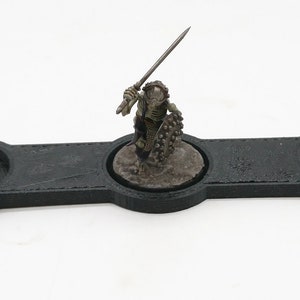 May include: A black plastic tray with three circular indentations for holding miniature figures. The tray is designed for tabletop gaming and features three miniature figures in various poses.
