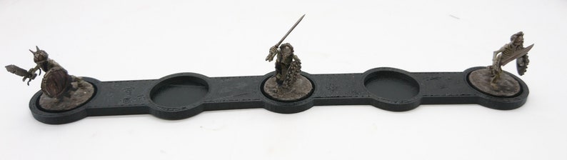 May include: A black plastic tray with three circular indentations for holding miniature figures. The tray is designed for tabletop gaming and features three miniature figures in various poses.
