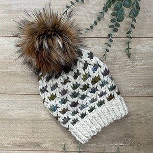 May include: A white knitted beanie with a brown and black faux fur pom pom. The beanie has a textured pattern with colorful accents.