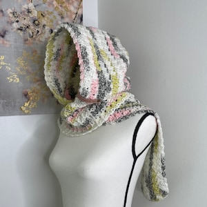 May include: A white, gray, pink, and green striped crocheted hooded scarf. The scarf is displayed on a white mannequin.