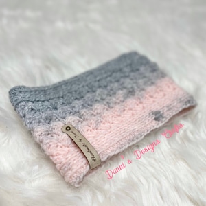 May include: A pink and grey knitted headband with a leather tag that says "Handmade by Dannie". The tag is attached to the headband with a metal rivet. The headband is on a white fluffy surface. Dannie's Designs Crafts is written on the bottom right corner of the image.