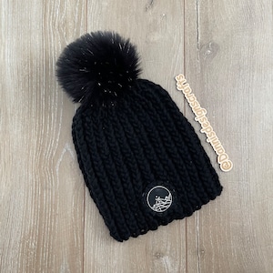 May include: Black knitted beanie with a fluffy pom pom and a small mountain logo patch. The beanie is on a woodgrain background. @outsideisnotsotscary