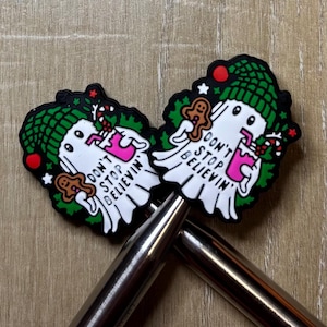 Puede incluir: Dos amuletos blancos con forma de fantasma con sombreros verdes y el texto "DON'T STOP BELIEVIN'". Cada amuleto sostiene un hombre de jengibre y una bebida rosa. Los amuletos están sobre una superficie de madera.