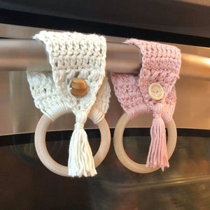 May include: Two crocheted oven mitt holders with wooden rings. One is white with a wooden button and the other is pink with a fabric button. Both have tassels.