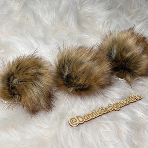 May include: Three brown faux fur pom poms on a white fluffy surface. The pom poms are soft and fluffy and would be great for adding a touch of texture to a hat or scarf.