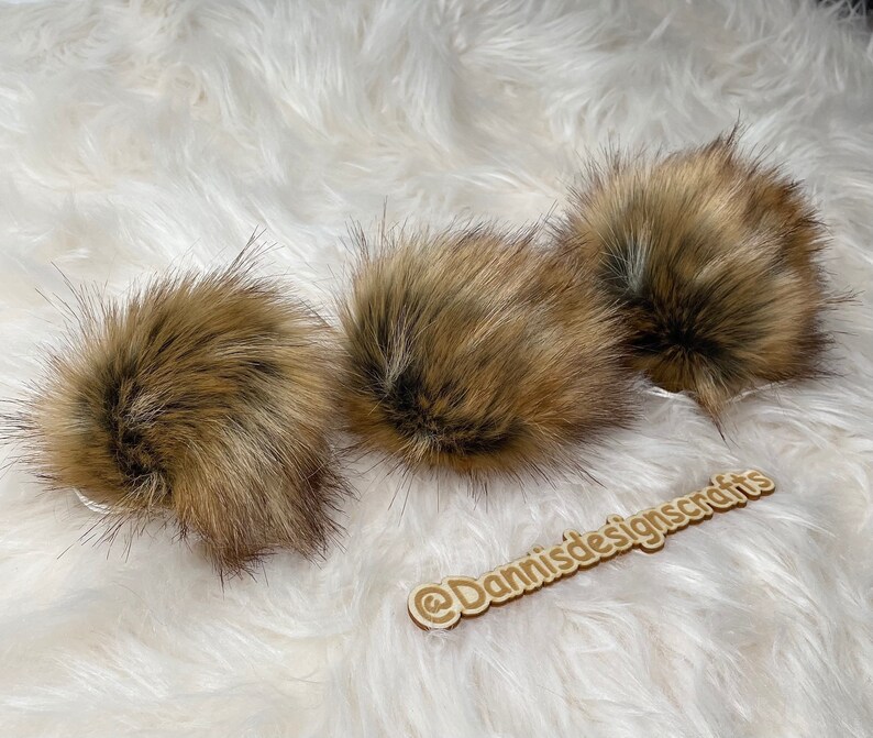 May include: Three brown faux fur pom poms on a white fluffy surface. The pom poms are soft and fluffy and would be great for adding a touch of texture to a hat or scarf.