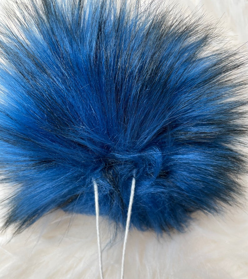 Peut inclure: Un pompon en fausse fourrure bleu et noir avec deux cordons blancs attach&eacute;s.
