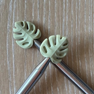 May include: Two light green monstera leaf-shaped stitch markers rest on the tips of two silver knitting needles. The needles are angled towards each other, creating a V-shape. The needles are marked with the text "Chicago 11 (8.0mm)".