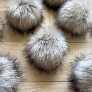 Könnte beinhalten: Eine Sammlung flauschiger Kunstfell-Bommel in Grau- und Brauntönen. Die Bommel sind rund und haben ein dichtes, strukturiertes Aussehen. Sie sind auf einer hellbraunen Holzoberfläche verstreut und erzeugen eine weiche, taktile Ästhetik.