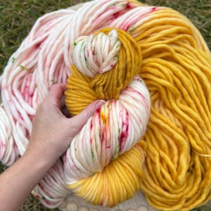 Op de afbeelding: Een close-up van kleurrijke garenstrengen. Het garen is een mix van wit met rode en groene spikkels en effen geel. Het garen is dik en grof, perfect voor brei- of haakprojecten. Het garen ligt op een houten basis.