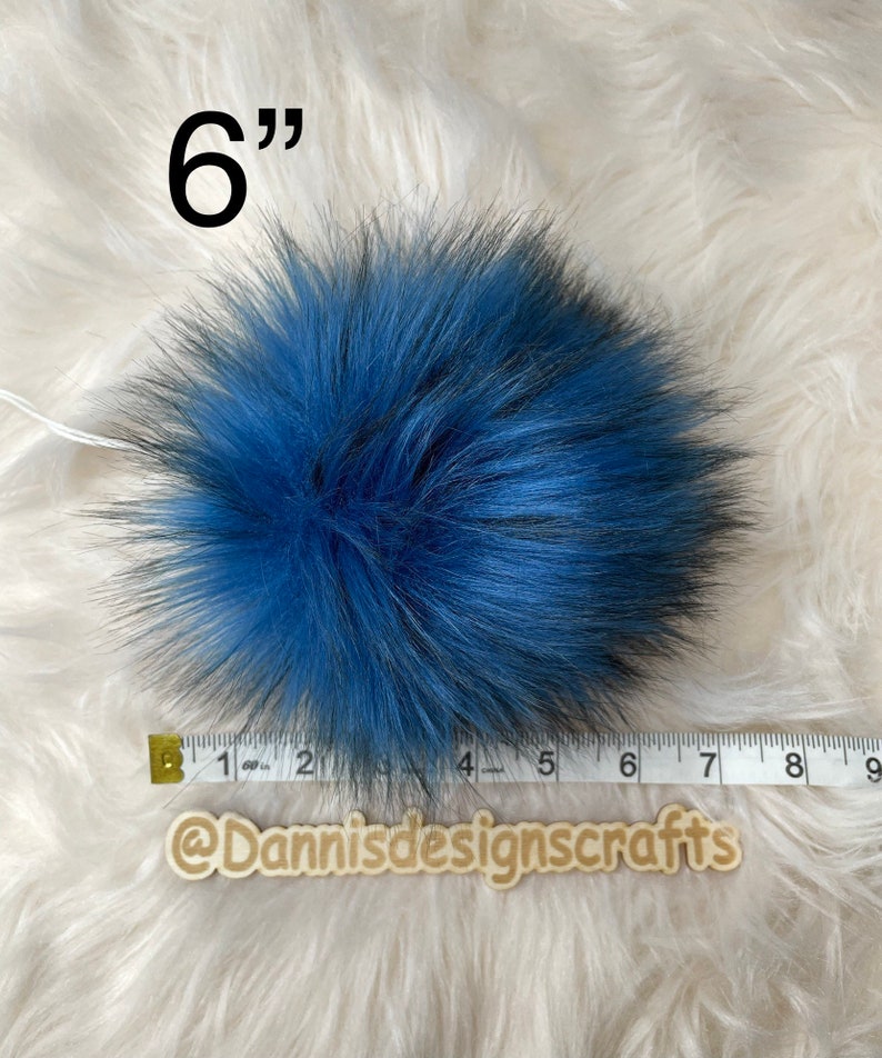 Peut inclure: Un pompon en fausse fourrure bleu et noir de 15 cm de diam&egrave;tre. Le pompon est pr&eacute;sent&eacute; sur une surface moelleuse blanche avec un ruban de mesure.