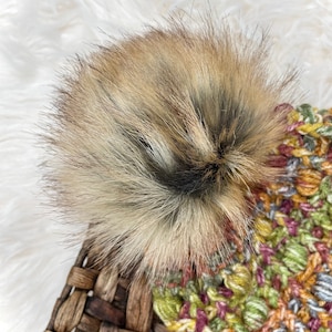 May include: A colorful crocheted beanie with a fluffy brown pom pom. The beanie is made with a variety of shades of green, yellow, orange, pink, and gray yarn.