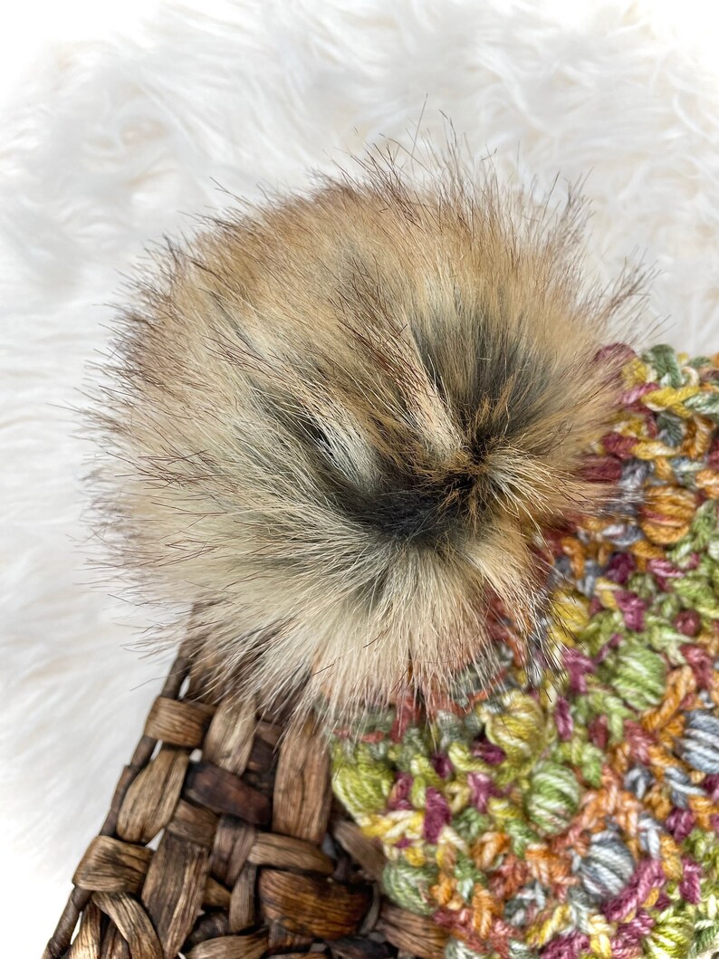 May include: A colorful crocheted beanie with a fluffy brown pom pom. The beanie is made with a variety of shades of green, yellow, orange, pink, and gray yarn.