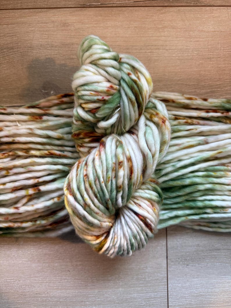 May include: A skein of thick, chunky yarn featuring a variegated color palette of cream, sage green, and burnt orange speckles. The yarn is twisted into a figure-eight shape, showcasing a soft, bulky texture with consistent ply throughout the entire length.