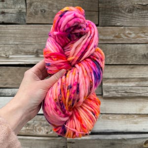 Könnte beinhalten: Ein lebendiges Knäuel grober Wolle in Rosa-, Orange- und Lilatönen. Die dicke, handgefärbte Wolle ist eng gewickelt und zeigt eine Mischung aus Farben. Die Wolle wird in einer Hand gehalten, mit einem Hintergrund aus Holzplanken.