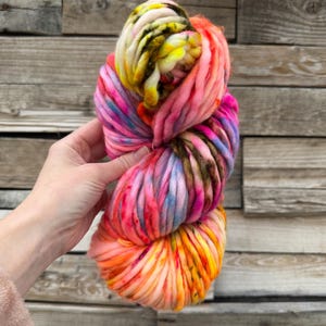 May include: A vibrant skein of super chunky yarn in a rainbow of colors. The yarn features shades of pink, orange, yellow, blue, and brown. The thick yarn is held in a person's hand against a wooden background.