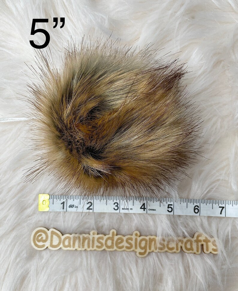 May include: A 5 inch brown and gray faux fur pom pom. The pom pom is measured with a white tape measure.