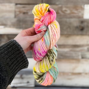 Puede incluir: Un ovillo de lana colorida en tonos rosa, amarillo, verde y melocotón. La lana gruesa se sostiene en una mano, mostrando sus vibrantes colores variados. La lana parece suave y gruesa, ideal para proyectos de tejido o ganchillo.