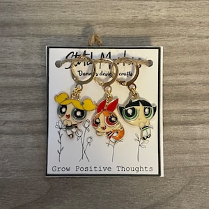 May include: Gold-tone hoop earrings featuring three cartoon characters with yellow, red, and black details. The earrings are attached to a white card with the text "Grow Positive Thoughts" and the brand name "Danny's design crafts".