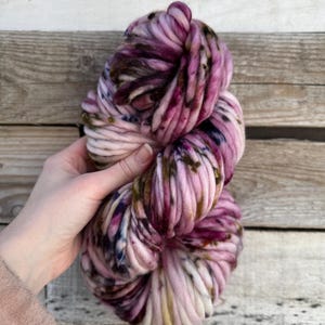 Pode incluir: Um grande novelo de fio grosso em tons de rosa, roxo e creme. O fio tem uma aparência espessa e texturizada com detalhes em roxo e marrom mais escuros. O fio é segurado contra um fundo de madeira.