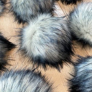 May include: Multiple fluffy faux fur pom poms in black and white. The pom poms are arranged on a wooden surface.
